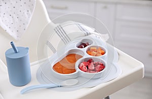 High chair with food in baby tableware on white tray indoors, closeup