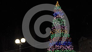 High Christmas tree on town square, decorated with garlands and colorful balloons in the night sky. little snow falls