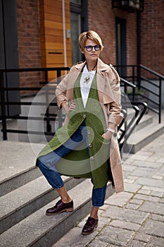 High Fashion Clothing. Beautiful Woman Wearing Fashionable Spring Or Fall Clothes beige trench coat, long green sweater,