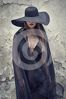 High Fashion Model in Black Hat hidden Face Grunge Wall Background. Stylish Woman Portrait with Red Lips and Necklace Jewelry.