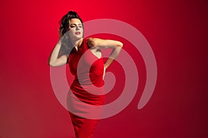 High fashion model wearing tight midi red dress and posing over gradient red-burgundy studio background in neon light.
