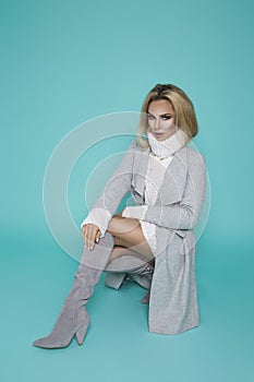 High fashion portrait of young elegant woman on blue background. Grey coat, white dress and elegant boots. Casual