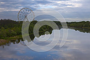 A high Ferris wheel stands on the bank of the river, in the water of which the evening sky is reflected