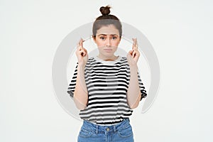 High hopes. Young woman cross fingers, makes wish, anticipates something, stands over white background