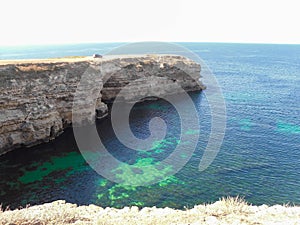 A high long rock that goes into the sea and a car and a man on top of the cliff. Sunny day, the sky and the clear turquoise water