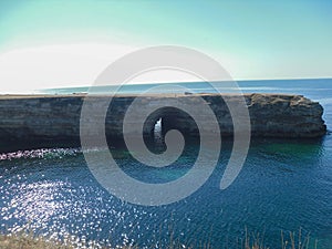 High long rock that goes into the sea and a tunnel inside it created by nature itself. A car and a man on top of a cliff. Sunny