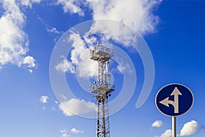 The high mast or pole with spotlights and a traffic sign `go straight or left turn` against a blue sky with white clouds, low a