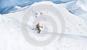 Svetlooranžová softshellová bunda vo vysokohorskom oblečení s trekingovými palicami zostupujúcimi zo zasneženého vrcholu hory. Aktívne