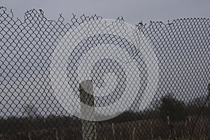 High net fence with barbed wire, concrete pillar, pole, beam against blue grey sky. meadow, field on background