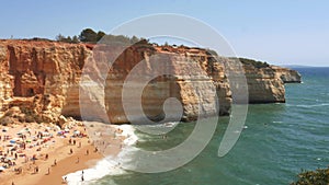 High perspective panning of gold-coloured cliffs, busy beach and turquoise ocean in Benagil beach, Lagoa, Algarve