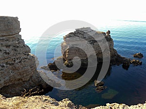 A high precipitous coastal rocky cliff and a small detached rock near the shore.