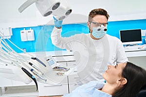 High-qualified dentist in uniform and magnifying dental loupes adjusting light lamp