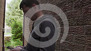 High quality 4k video of a young man who is texting messages on his cellphone standing in a shade of the red bricks wall.