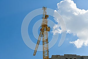 High-rise construction crane with a long arrow of yellow color against the blue sky over a new multi-storey building of concrete