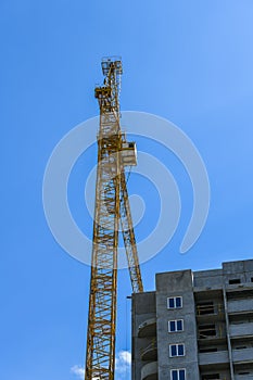 High-rise construction crane with a long arrow of yellow color against the blue sky over a new multi-storey building of concrete