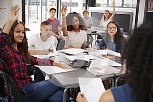 High school kids raise hands, teacher sitting at their desk