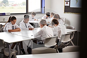 High School Students Wearing Uniform Sitting And Working Around Table In Lesson