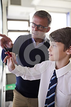 High School Teacher With Male Student Wearing Uniform Using Interactive Whiteboard During Lesson