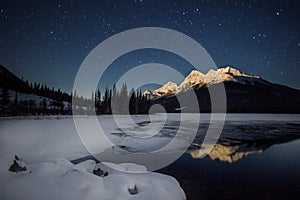 High snow covered mountain, in full moon light with a half frozen lake under night sky full of stars, Banff national Park, Canada