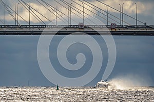 a high-speed passenger hydrofoil boat passes under a cable-stayed bridge in sunny weather, dark storm clouds in the