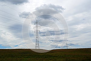 High tension electricity poles and cables over green grass field and gray cloudy sky in the countryside