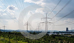 High voltage lines and power pylons in a green landscape, gray sky and chimney of a nuclear power plant in the background.