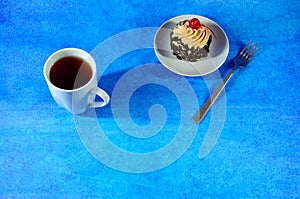 A high white mug with tea, next to a saucer with a biscuit cake in chocolate chips and cherry on a cream and a teaspoon. The whole