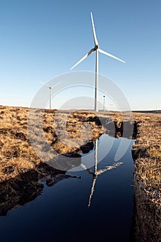 High wind power turbine in turf bog, reflection in water. Cloudy sky background. Renewable green energy production, source of