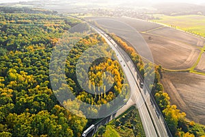 Highway between Autumn forest and cultivated ground with yellow trees at sunset in autumn. Aerial view of the traffic on speedway.