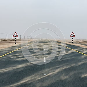 Highway road among desert during sand storm with road signs with camel warning