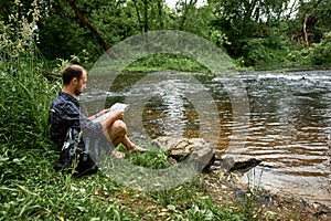 Hiker with a backpack and map sitting on the bank of a river and exploring of the area