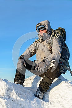 Hiker with backpack sitting and having rest on the top of a snow-covered rock over the winter mountains