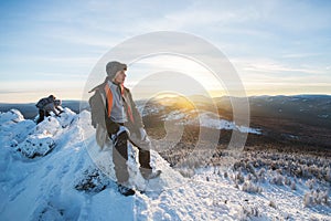 Hiker with backpack sitting and having rest on the top of a snow-covered rock over the winter mountains at beautiful