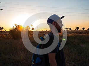 Hiker with backpack standing on a hill and enjoying sunset. Bearded man in snapback walking. Local travel nature concept