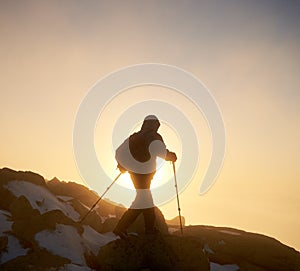 Hiker with backpack and trekking poles on rocky mountain on background of raising sun and misty sky.