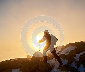 Hiker with backpack and trekking poles on rocky mountain on background of raising sun and misty sky.