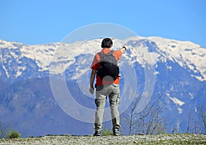 hiker with a black backpack on his shoulders pointing to the snow-capped mountains that will be his next destination