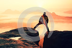 Hiker in black is jumping between the rocky peaks. Wonderful daybreak in rocky mountains, heavy orange mist in deep valley.