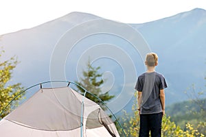 Hiker child boy resting standing near a camping tent at mountains campsite enjoying view of summer nature