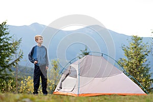 Hiker child boy resting standing near a camping tent at mountains campsite enjoying view of summer nature