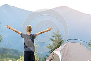 Hiker child boy standing near a tent in mountain campsite with raised hands enjoying view of nature