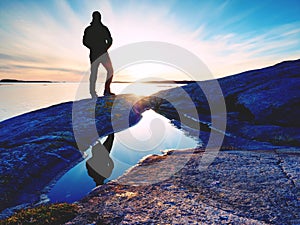 Hiker in dark sportswear with poles and sporty backpack. Coastline trail on rocky shore. Alone tourist enjoy