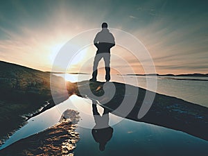 Hiker in dark sportswear with poles and sporty backpack. Coastline trail on rocky shore. Alone tourist enjoy