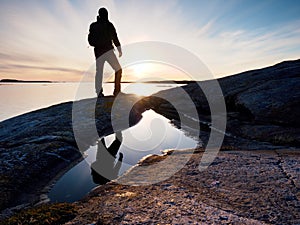 Hiker in dark sportswear with poles and sporty backpack. Coastline trail on rocky shore. Alone tourist enjoy