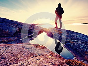 Hiker in dark sportswear with poles and sporty backpack. Coastline trail on rocky shore. Alone tourist enjoy