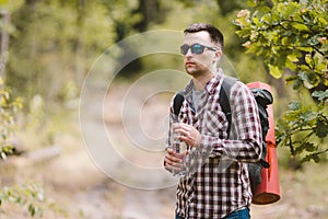 Hiker Drinking Water In Forest. Tired man drinking water from bottle in woodland. Handsome Traveler With Backpack And Flask In