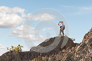 Hiker girl with backpack standing on top of mountain. Tourist looks from peak of hill to beautiful landscape on sky background