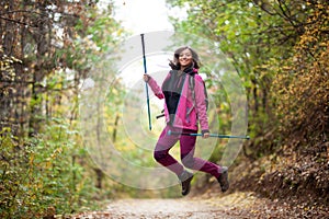 Hiker girl jumping on a trail in the mountains. Backpacker with hiking poles and pink jacket in a forest. Happy lifestyle outdoors