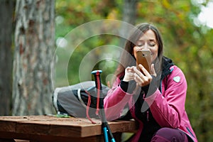 Hiker girl resting on a bench in the forest. Backpacker with pink jacket holding cell phone