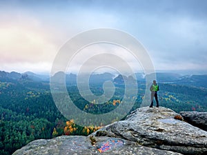 Hiker in green windcheater, cap and dark trekking trousers stand on mountain peak rock.